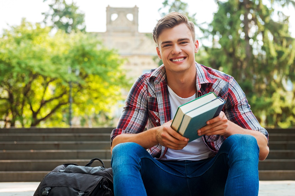 Male student holding textbooks and smiling while sitting at the outdoor staircase with university building in the background