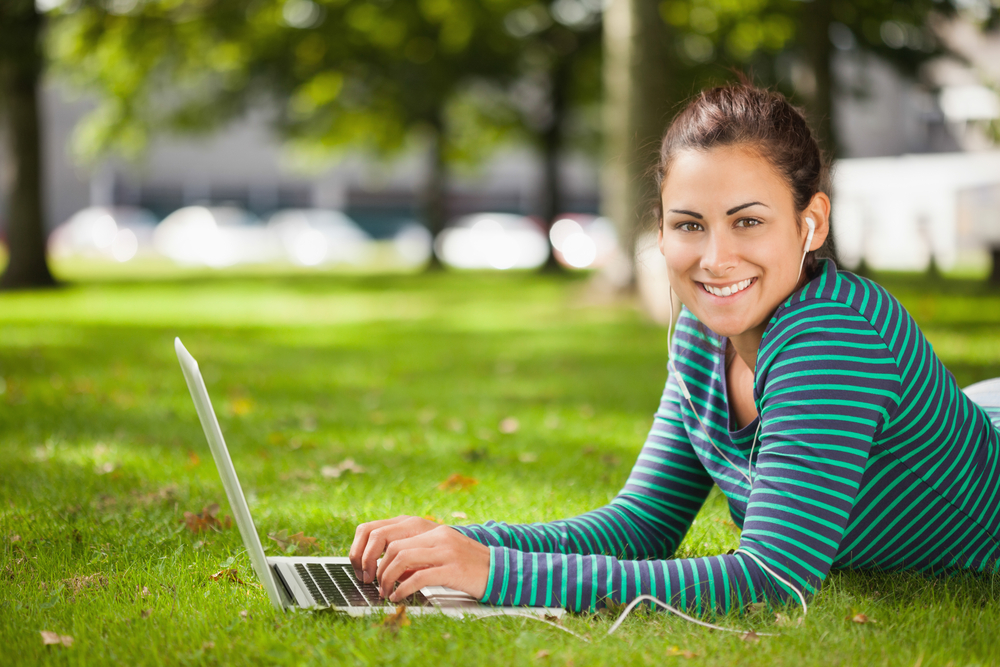 Student lying on grass using laptop looking at camera on campus at college